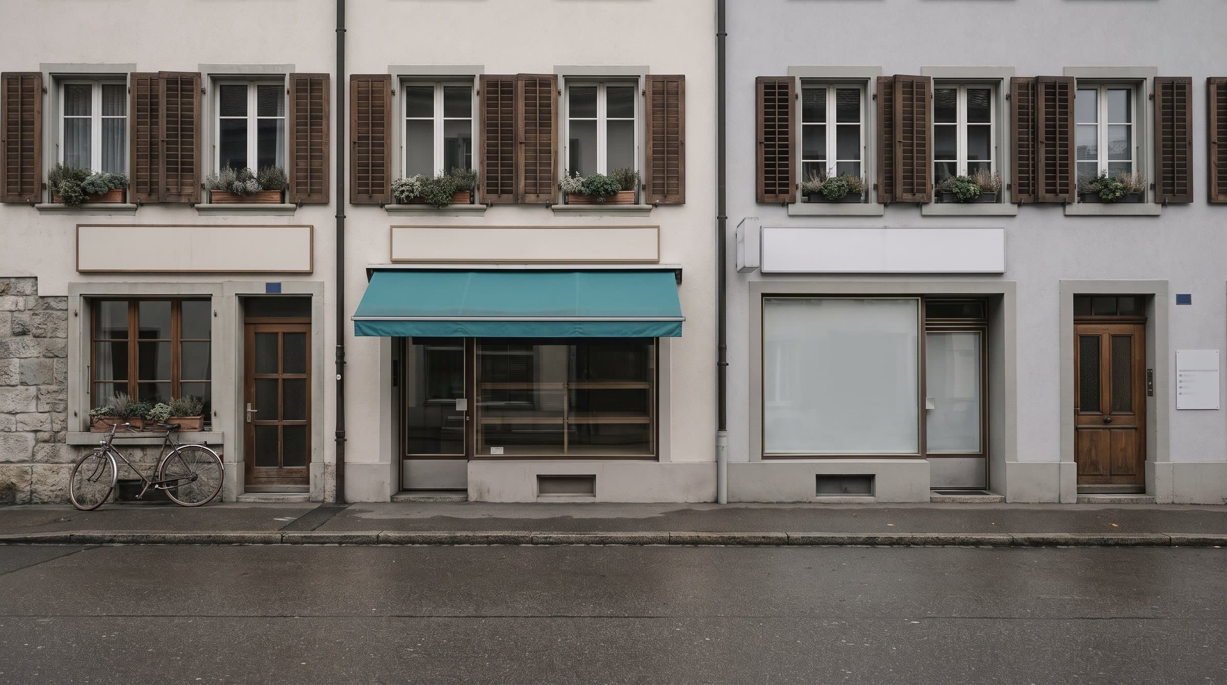 Ladenzeile in einem Schweizer Städtchen: drei kleine Geschäfte nebeneinander, verputzte Fassaden mit Holzläden, eine türkise Markise über einem Schaufenster, ein Velo an der Wand. Alle Schilder sind leer.